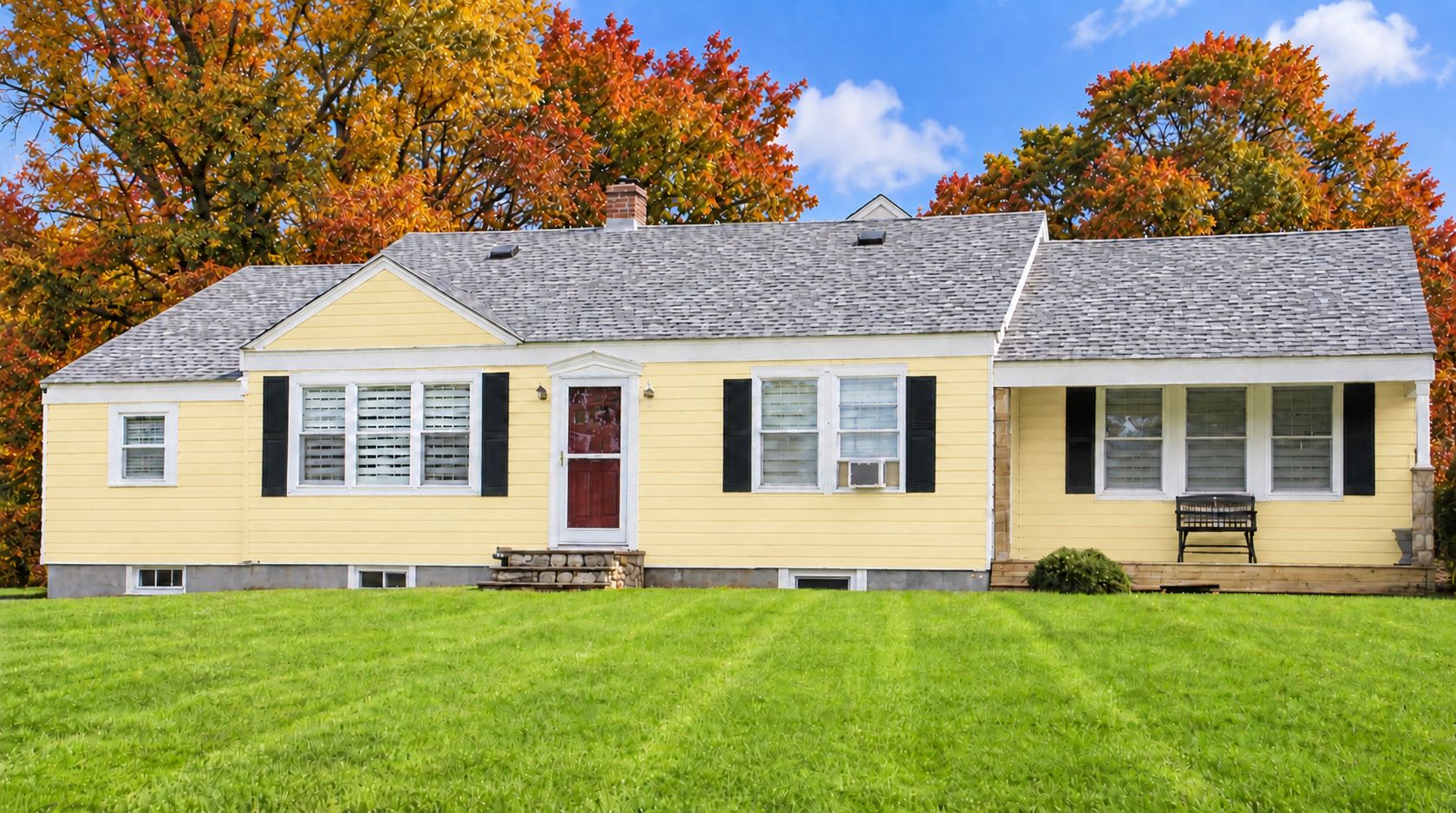 A two-story yellow house with a red door, white shutters, and a stone foundation.