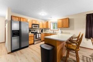 A modern kitchen with wooden cabinets, stainless steel appliances, a central island with bar stools, and a window providing natural light. - Spacious 2BR | Near Downtown & Sanford | Garage (Sioux Falls)
