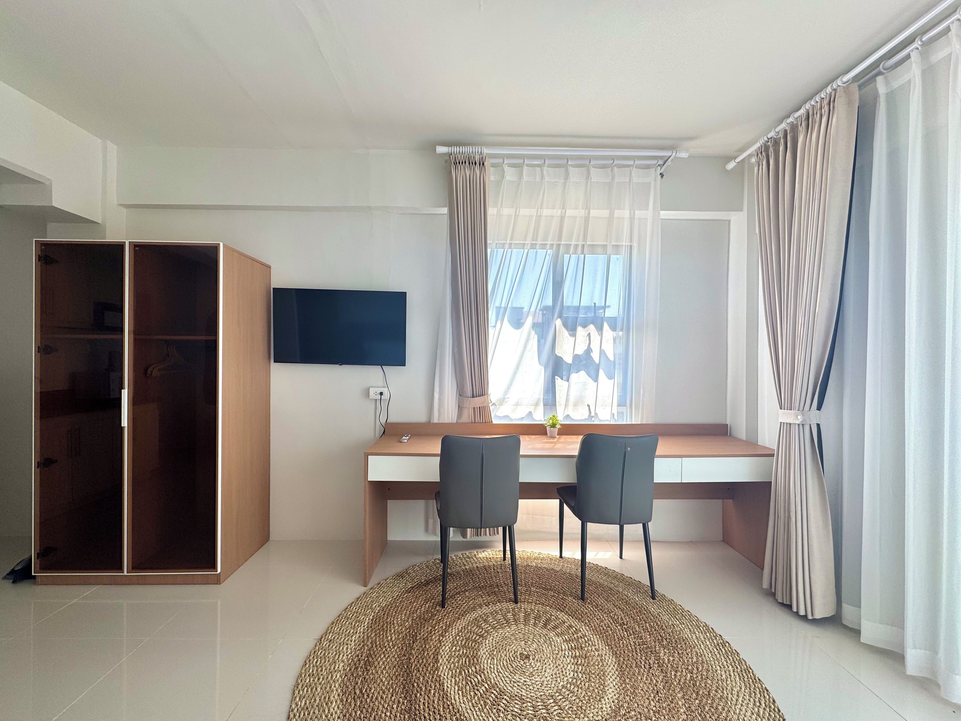 A room with a wooden desk, two chairs, a television, a wardrobe, and a window with curtains.