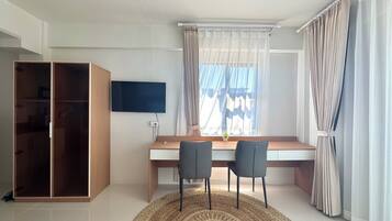 A room with a wooden desk, two chairs, a television, a wardrobe, and a window with curtains.