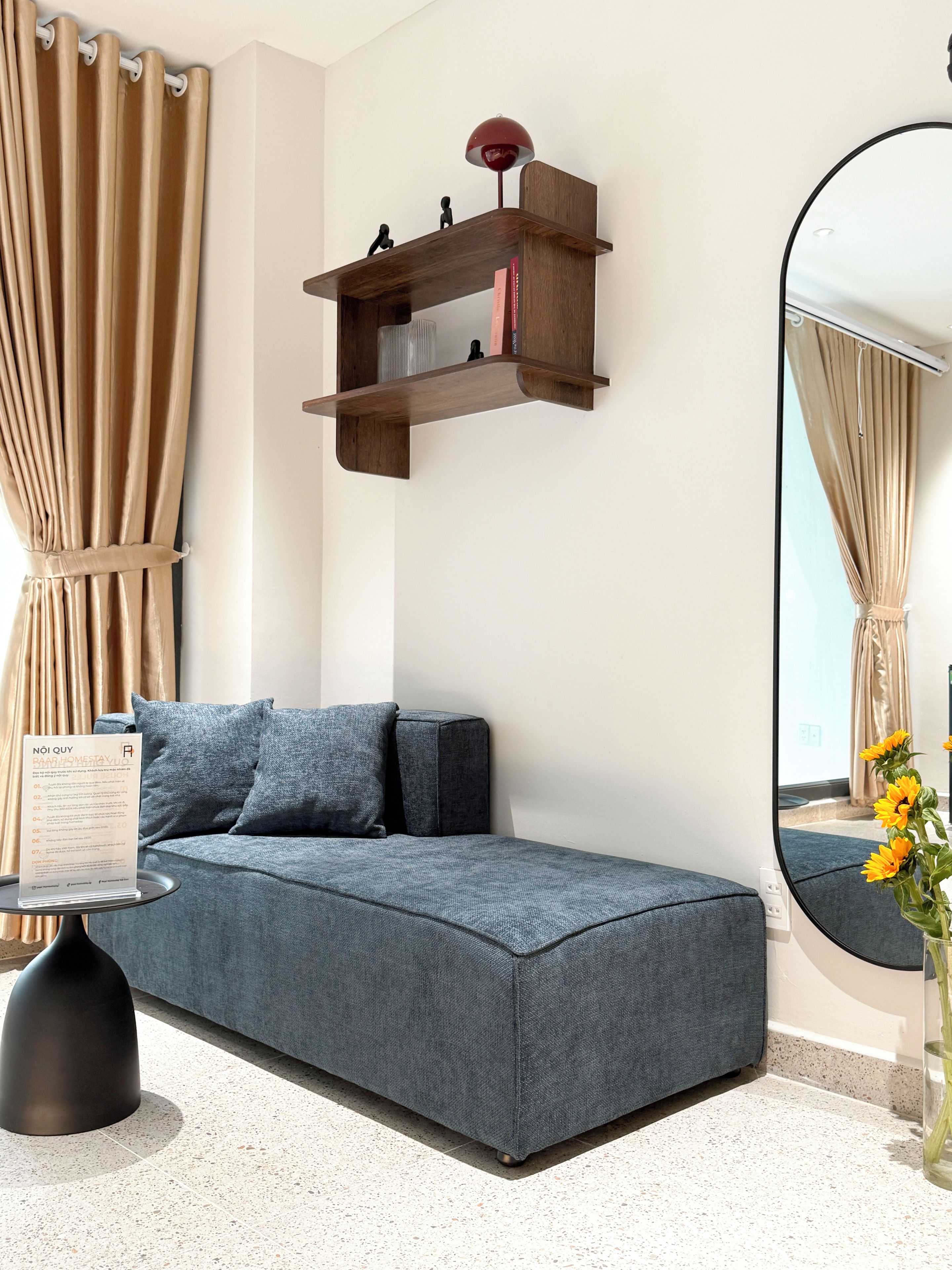 A modern living room with a grey sofa, a small wooden shelf with books, a round mirror, and a vase with yellow flowers.
