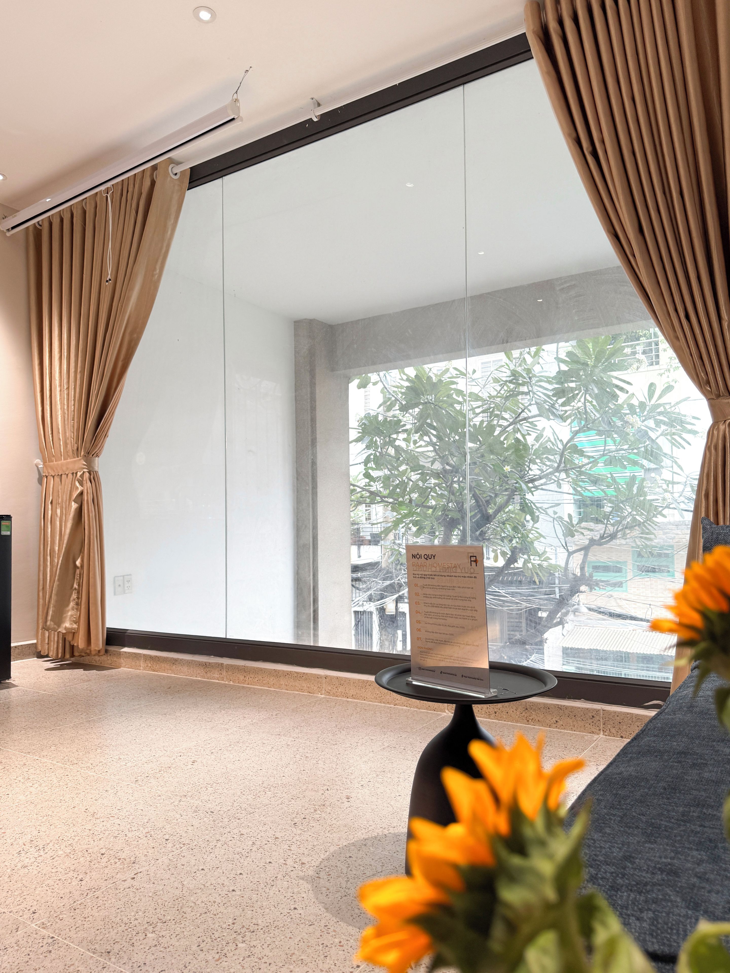 A hotel room with a large window, curtains, a table with a sign, and a flower in the foreground.