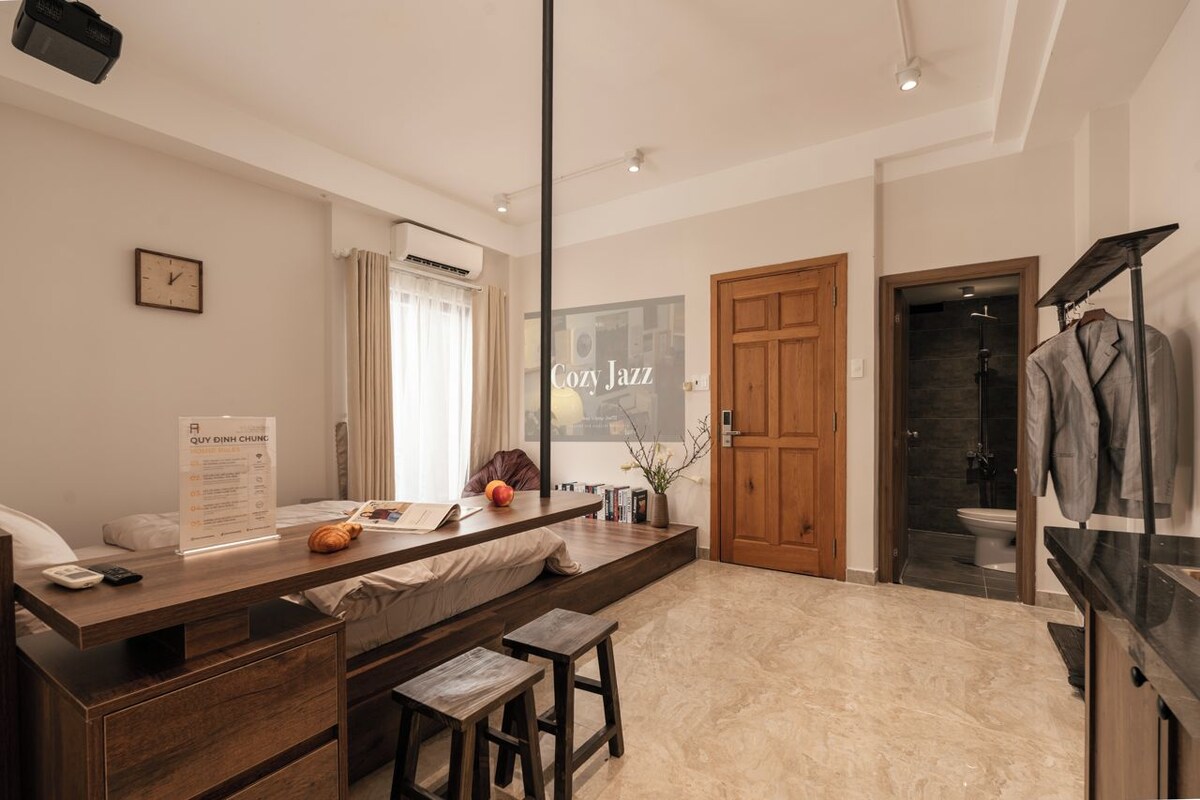 Une chambre d’hôtel moderne avec un bureau en bois, un lit, un petit coin salon, une salle de bain avec douche et une vue sur la ville.