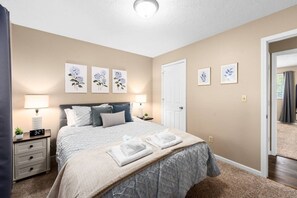 A bedroom with a bed, bedside lamps, a nightstand, a door, and wall art. - Pioneer Place | King Bed | Deck | 3-Min to Avera (Sioux Falls)