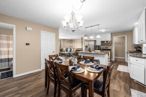 A modern kitchen with a central island, a dining area with wooden chairs and a table set for four, and a chandelier above the dining table. - Pioneer Place | King Bed | Deck | 3-Min to Avera (Sioux Falls)