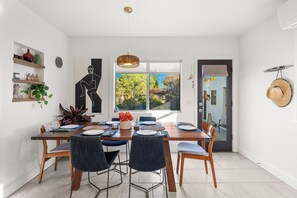A dining area with a wooden table set for four, surrounded by chairs, a hanging pendant light, and a view of a garden through a window. - SAN DIEGO SPA VILLA - Cozy Yard, Firepit, Rec Room (San Diego)