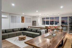 A modern living room with a sectional sofa, a wooden dining table, and a kitchen in the background. - The Bunkhouse - Bicheno Accommodation (Bicheno)