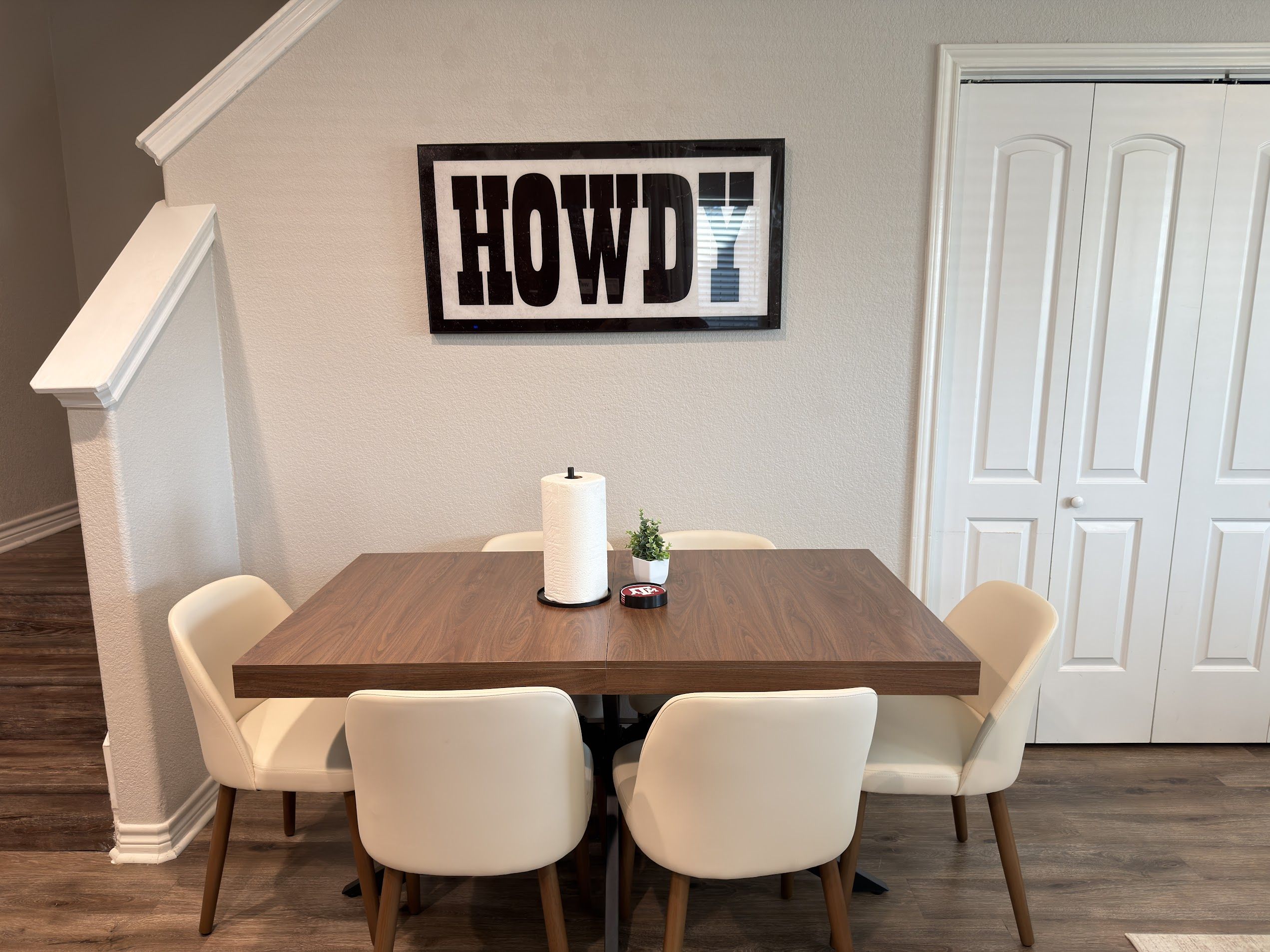 A dining area with a wooden table, four white chairs, a wall-mounted sign, and a door in the background.