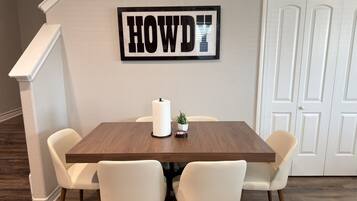 A dining area with a wooden table, four white chairs, a wall-mounted sign, and a door in the background.