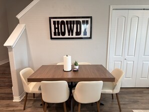 A dining area with a wooden table, four white chairs, a wall-mounted sign, and a door in the background.