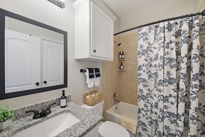 Een badkamer met een wastafel met een granieten blad, een wit toilet, een douche met een bloemenprint gordijn en een spiegel met een zwarte lijst.