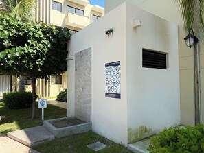 A small, white building with a shower area, a sign with symbols, and a palm tree nearby. - Relax at Oceanna | Rooftop, Jacuzzi, and 3 Pools (Mazatlán)