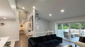 A modern living room with a black sofa, a dining area with a white table, and a staircase leading to a balcony with outdoor furniture.
