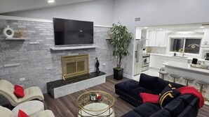A modern living room with a fireplace, a flat-screen TV, and a kitchen in the background. - Catskills Stone Getaway - 6BR, Sleeps 14, Game Room, Firepit, Near Skiing (Purling)