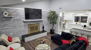 A modern living room with a fireplace, a flat-screen TV, and a kitchen in the background. - Catskills Stone Getaway - 6BR, Sleeps 14, Game Room, Firepit, Near Skiing (Purling)