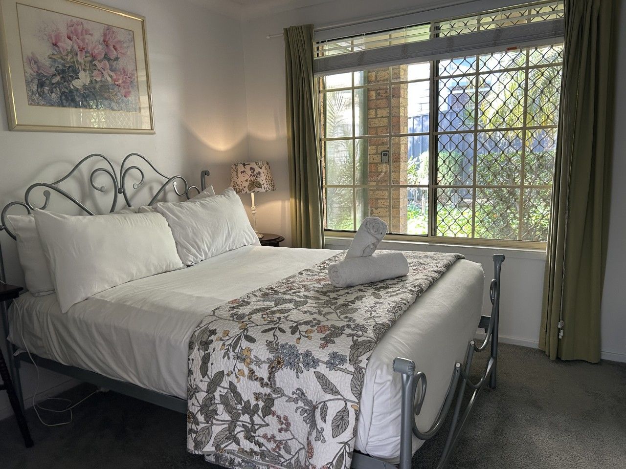 A neatly made bed with a floral bedspread and white pillows, a metal-framed headboard, a bedside table with a lamp, and a window with a view of greenery outside.