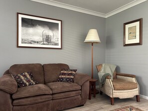 Un salon avec un canapé marron, un fauteuil à motifs, une lampe sur pied et deux tableaux encadrés accrochés au mur.