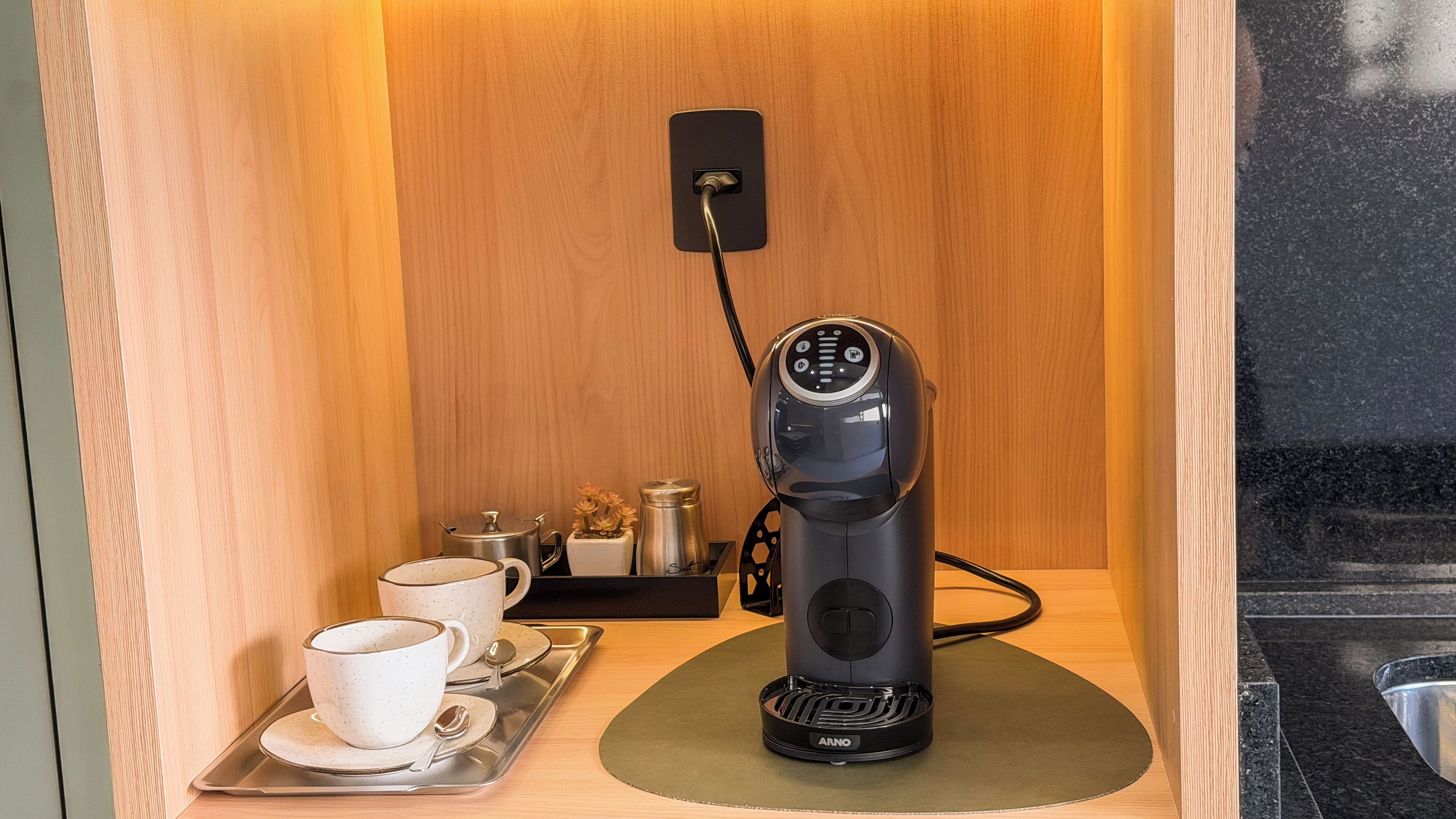 Quarto compacto com máquina de café, bandeja com chávenas e colher, e uma pequena mesa com chaleira e saleiro e pimenteiro.