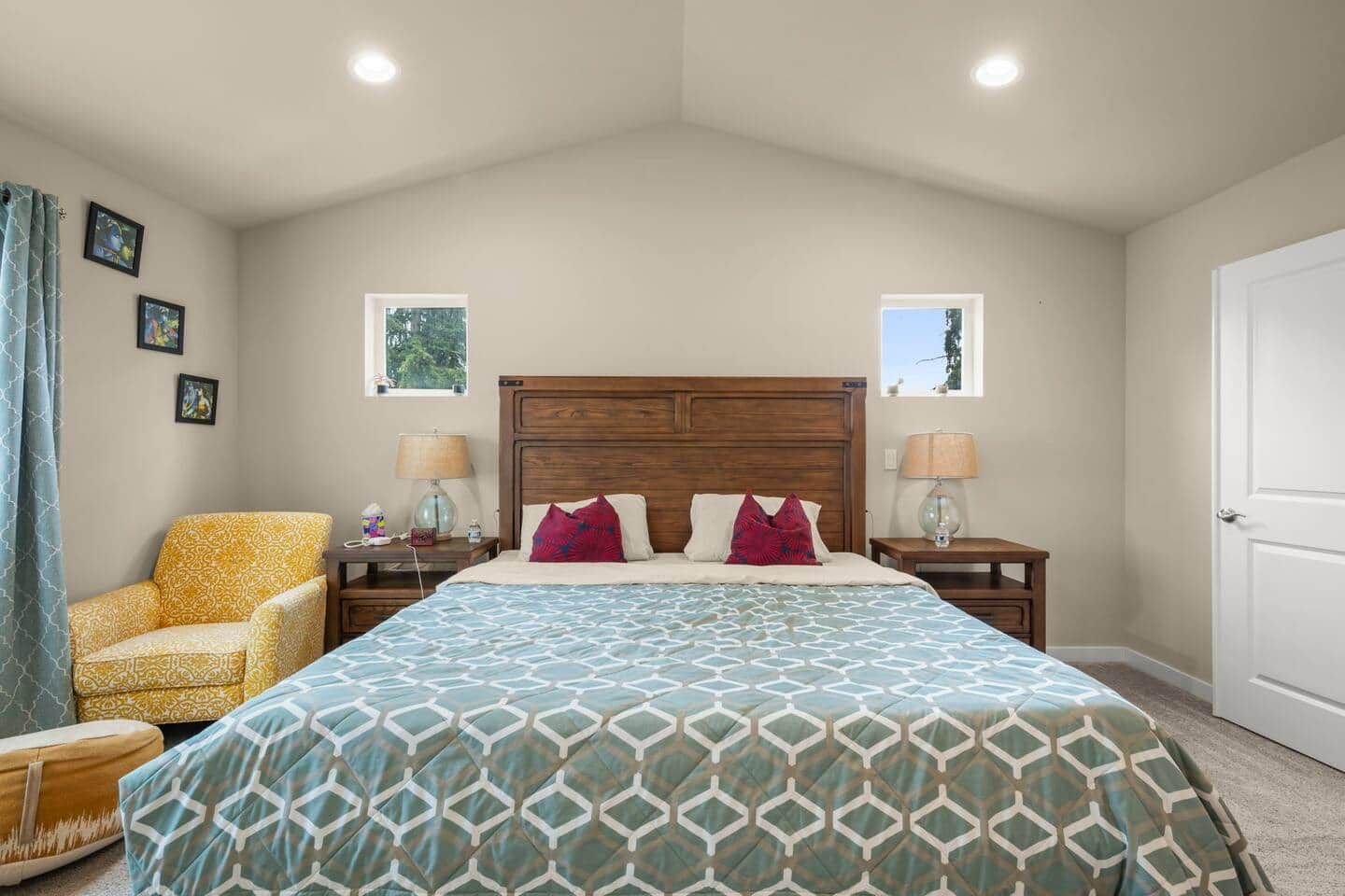 Un dormitorio con una cama grande, dos mesitas de noche, una silla y dos ventanas.