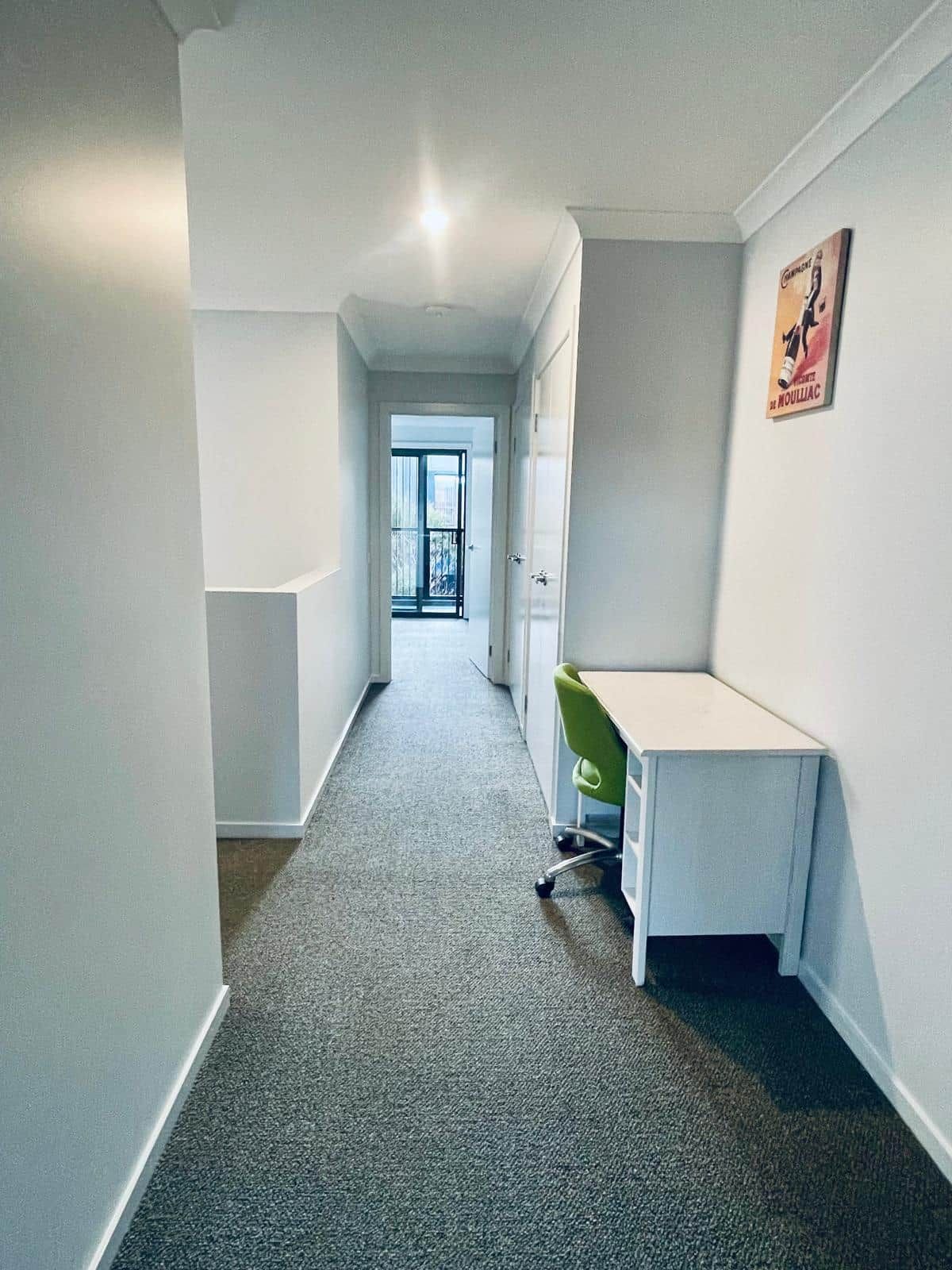 Een smalle gang met een tapijtvloer, een wit bureau met een groene stoel, en een deur die leidt naar een balkon.