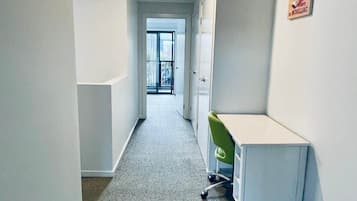 Een smalle gang met een tapijtvloer, een wit bureau met een groene stoel, en een deur die leidt naar een balkon.