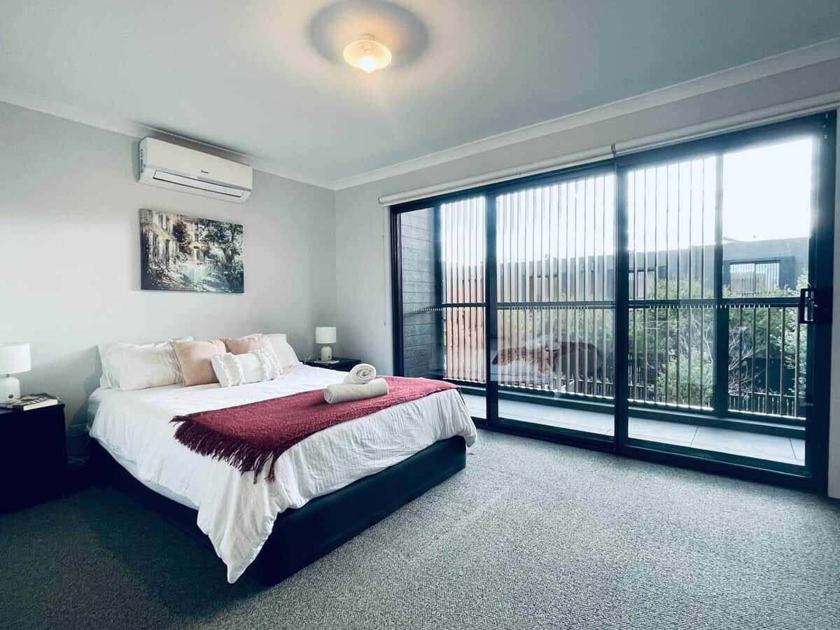 A bedroom with a large bed, a nightstand, a wall-mounted air conditioner, a window with blinds, and a view of the outdoors.