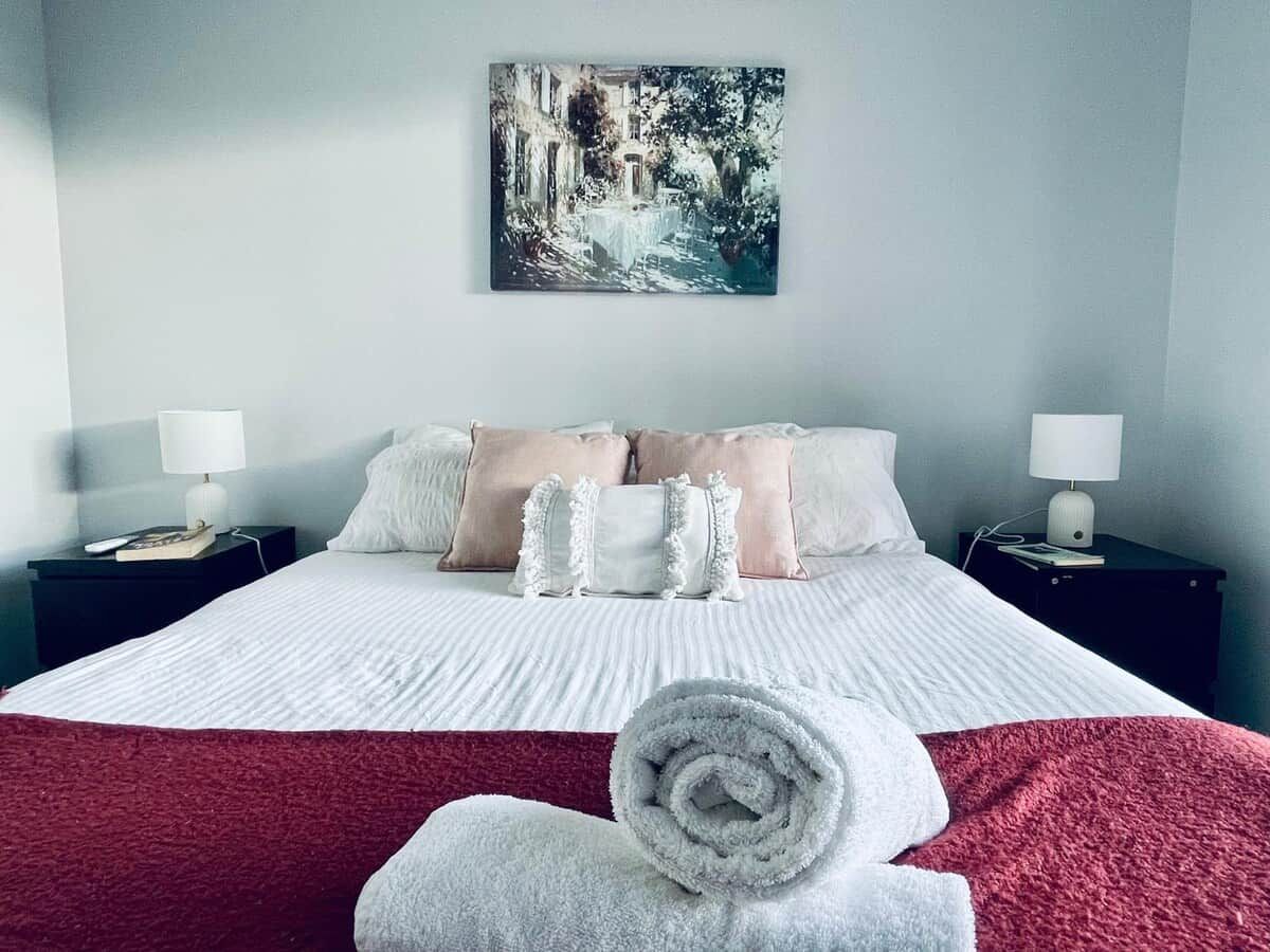 A neatly made bed with a red blanket, white pillows, and a rolled towel. There is a painting on the wall above the bed.