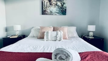 A neatly made bed with a red blanket, white pillows, and a rolled towel. There is a painting on the wall above the bed.