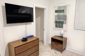 Ein Zimmer mit einem wandmontierten Flachbildfernseher, einem Holzkontenmöbel mit Schubladen, einem kleinen Nachttisch mit Lampe und einem dekorativen Wandteppich.