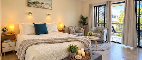 A bedroom with a bed, bedside tables, a nightstand, a chair, a small table, a window with curtains, and a balcony.