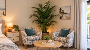 Quarto com cama, duas poltronas estampadas, mesa de centro redonda, planta em vaso e mesa de cabeceira com abajur.