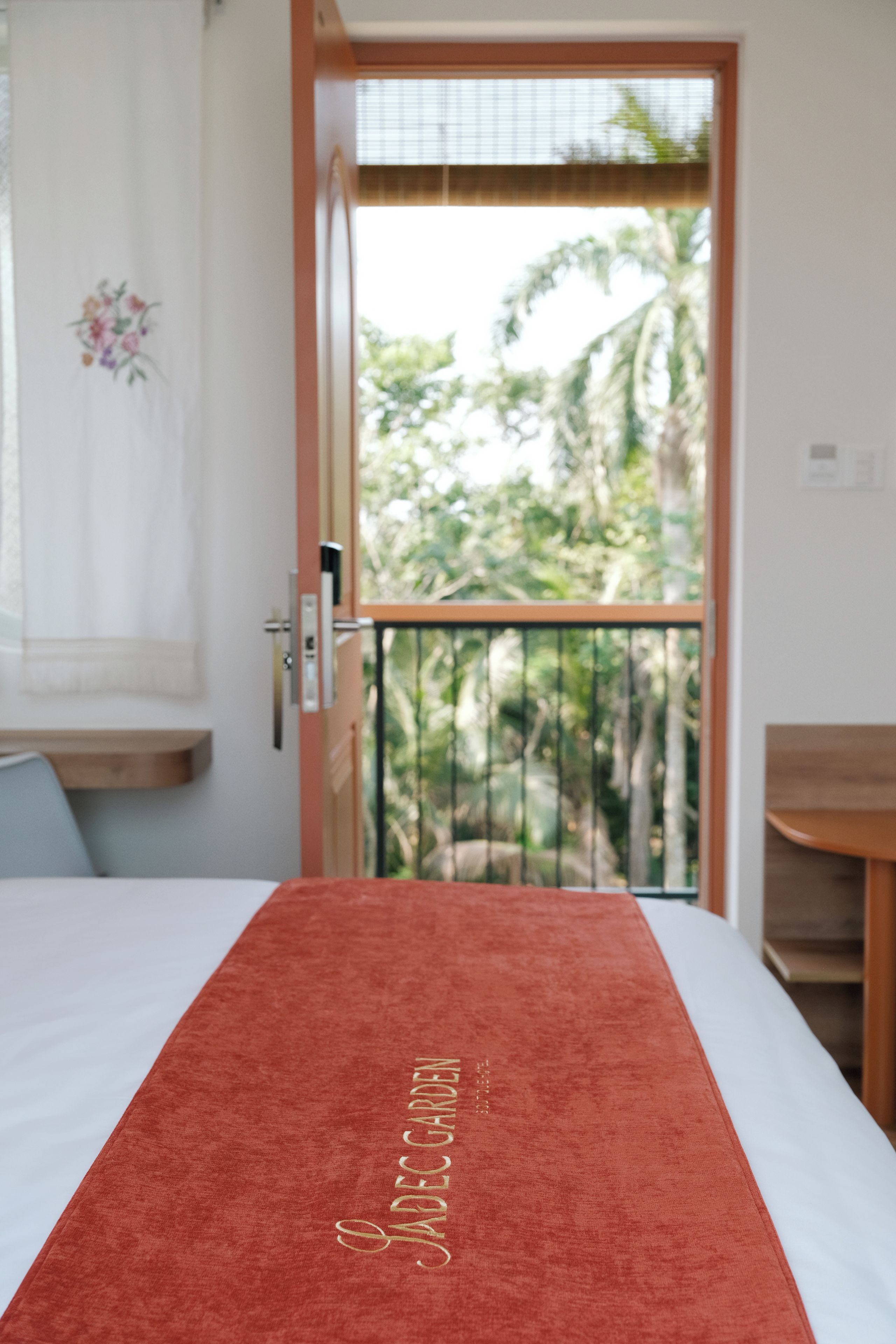 A room with a bed, a wooden door leading to a balcony, and a view of greenery outside.