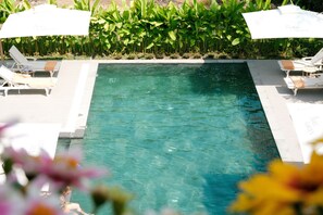 Una piscina rectangular con agua cristalina, rodeada de sillones de descanso blancos y sombrillas, y una exuberante franja de plantas verdes.