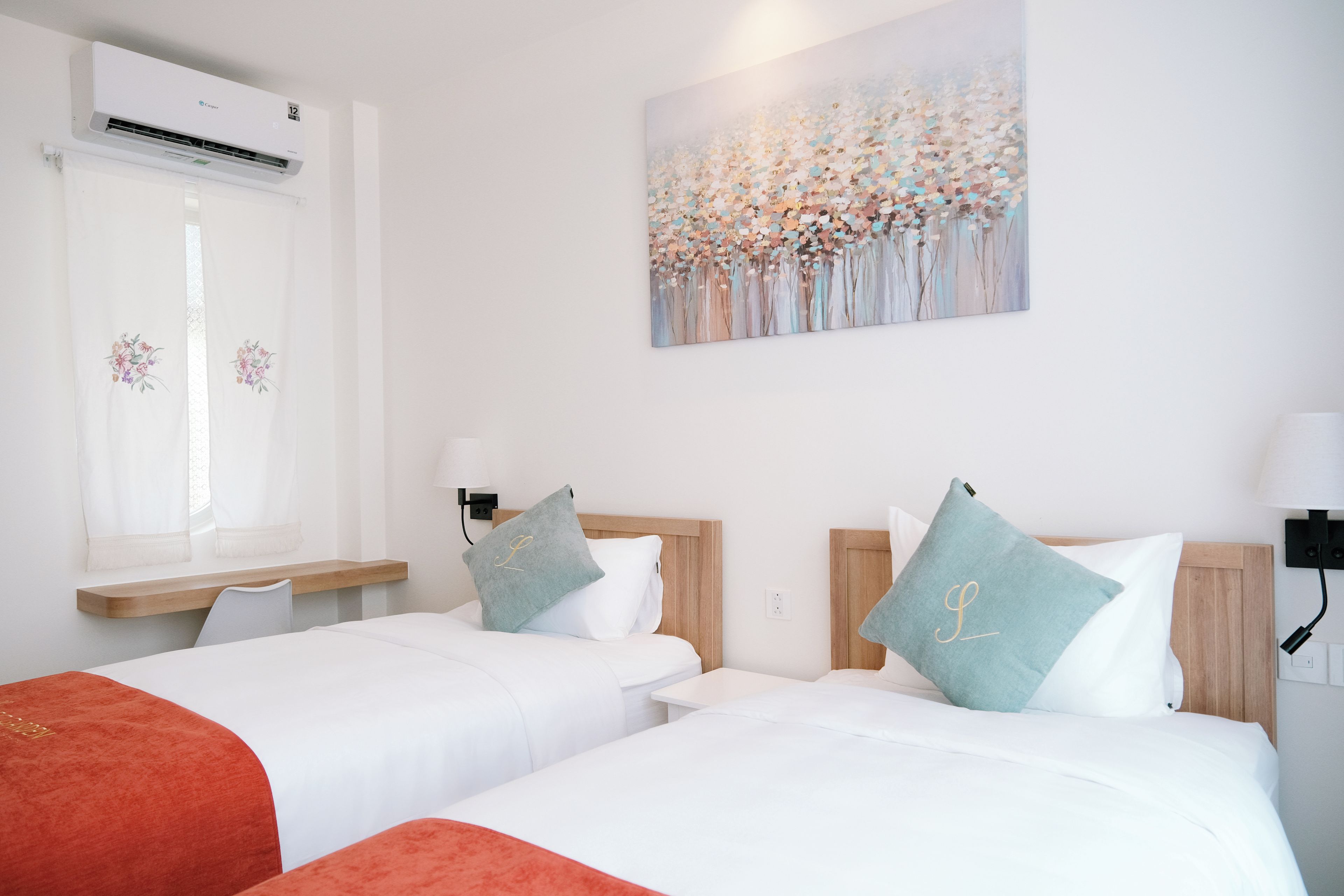 A hotel room with two beds, a window with curtains, an air conditioning unit, a painting on the wall, and a bedside table with a lamp.