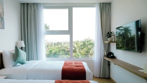 A hotel room with two beds, a large window with a view of greenery, a mounted TV, and a bedside table with a lamp.