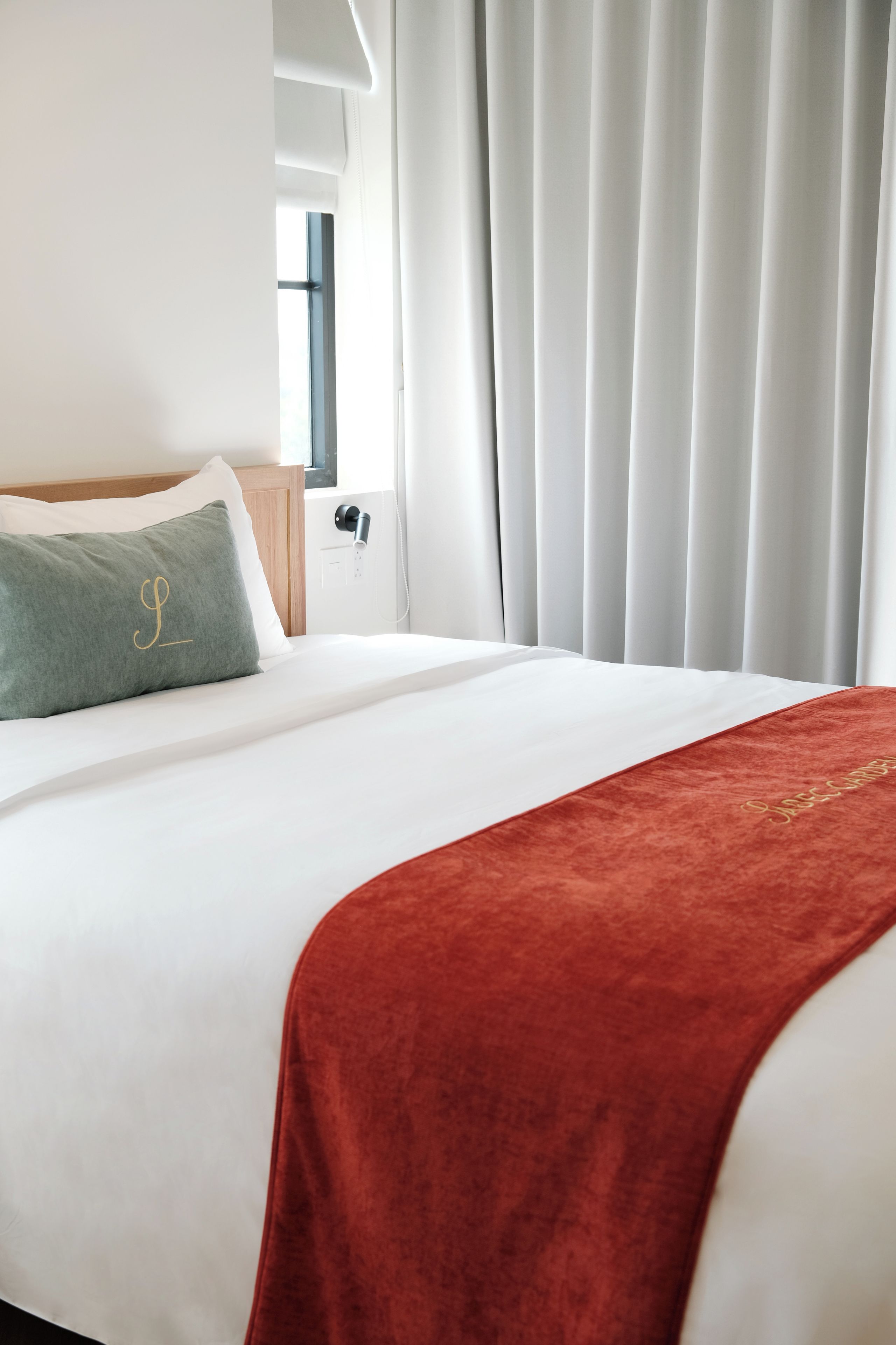 A hotel room with a neatly made bed, a red throw blanket, and a green pillow with a monogram.