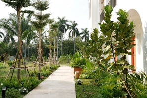 A pathway lined with various plants and trees, leading to a building with a white facade and an arched entrance.