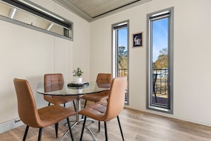 A dining area with a glass table and four chairs, large windows, and a view of the outdoors.