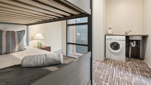 A compact hotel room with a bed, a nightstand, a lamp, a window, and a washing machine.