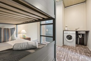 A compact hotel room with a bed, a nightstand, a lamp, a window, and a washing machine.