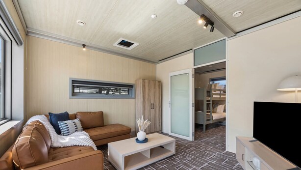 A modern living room with a sofa, coffee table, and a television. There is a sliding door leading to a bedroom with a bunk bed.