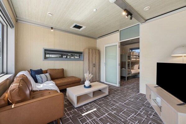 A modern living room with a sofa, coffee table, and a television. There is a sliding door leading to a bedroom with a bunk bed.