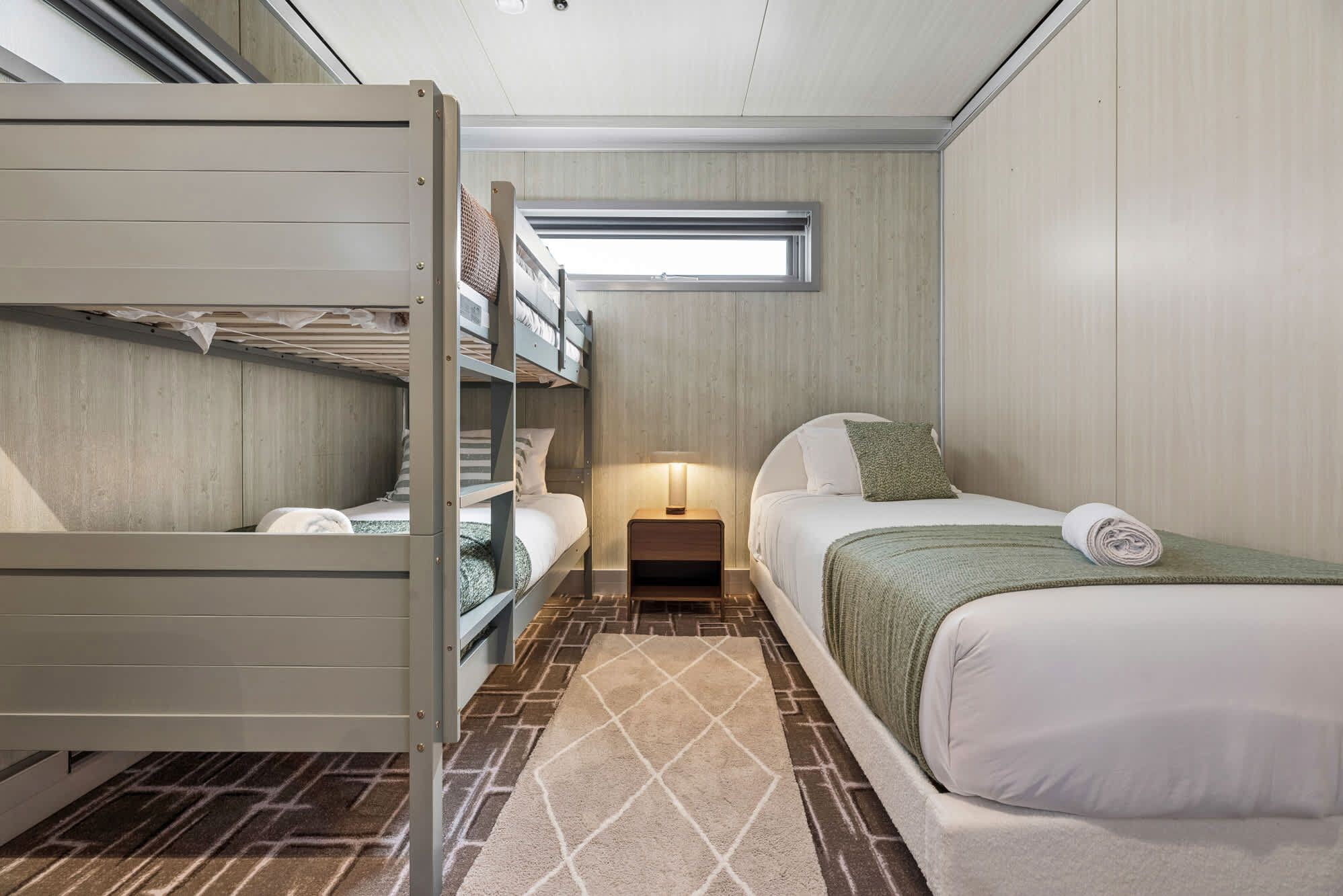 A compact hotel room with bunk beds, a single bed, a nightstand, and a window.