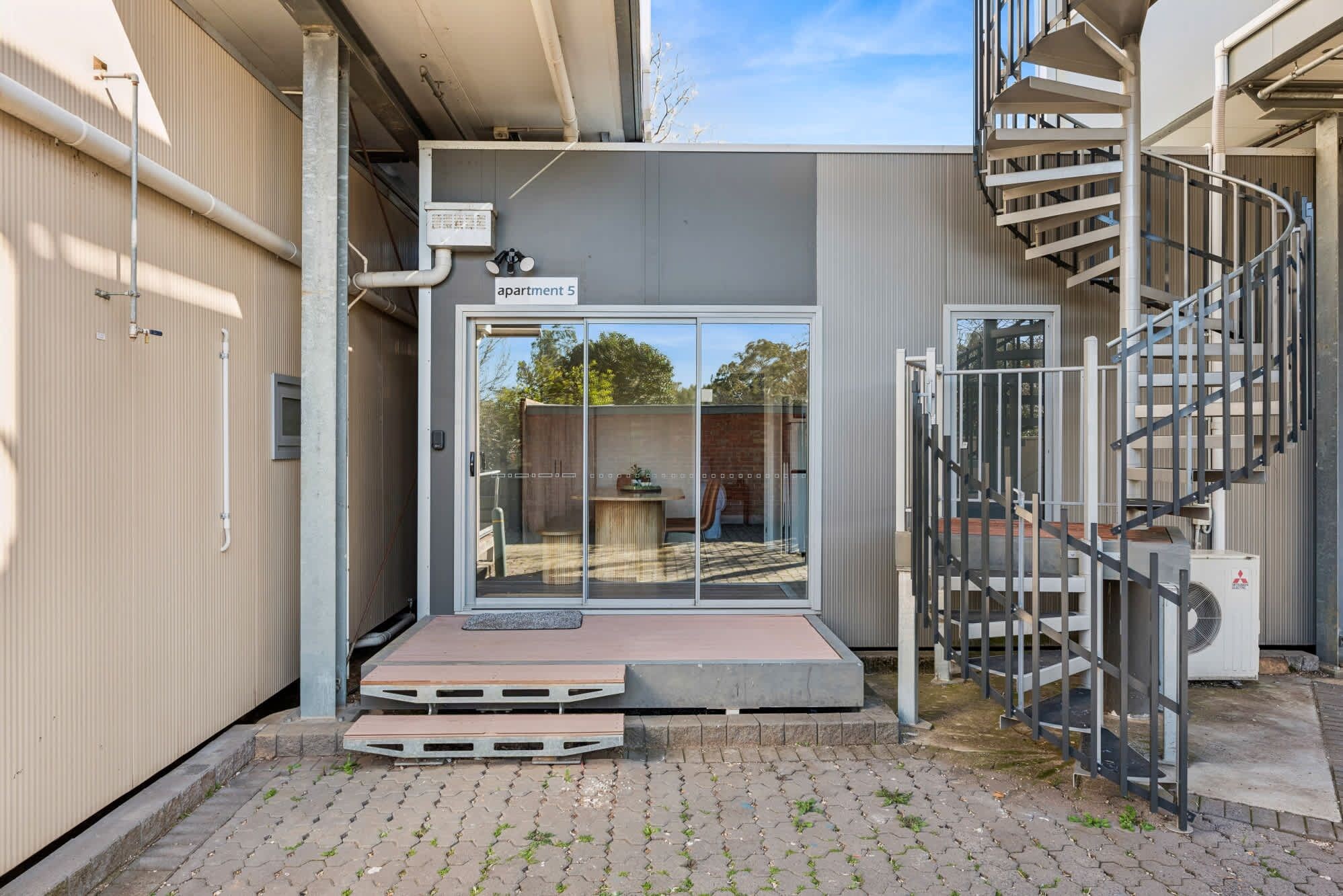 A modern apartment with a glass door entrance, a small outdoor seating area, and a spiral staircase.