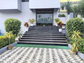 Degraus externos levam a uma entrada de edifício, cercados por plantas em vasos e um pavimento estampado.