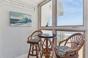 A room with a large window, wicker chairs, a small round table, and a painting of a wave. - Direct Oceanfront Condo in New Smyrna Beach on No-Drive Beach, 2B2B (New Smyrna Beach)