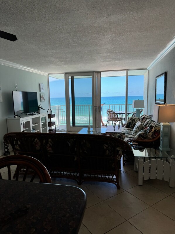 A living room with a television, a sofa, and a dining table. There is a balcony with a view of the ocean.