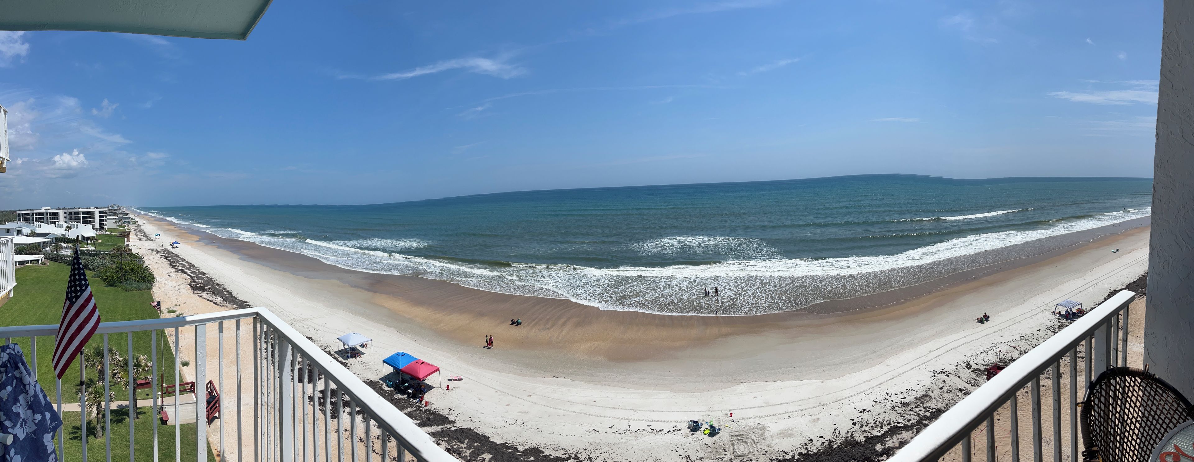 A beach view from a balcony with a railing, a flag, and beach umbrellas.