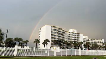 A multi-story hotel with a white facade and yellow trim, surrounded by a white picket fence and palm trees.