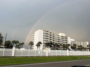 A multi-story hotel with a white facade and yellow trim, surrounded by a white picket fence and palm trees.